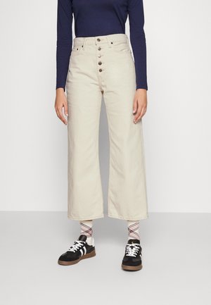 Pilkos plačios kojos džinsai su aukštu juosmeniu, priekyje užsegami mygtuku ir minkšta tekstūra, derinami su juodais sportbačiais ir raštuotomis kojinėmis.