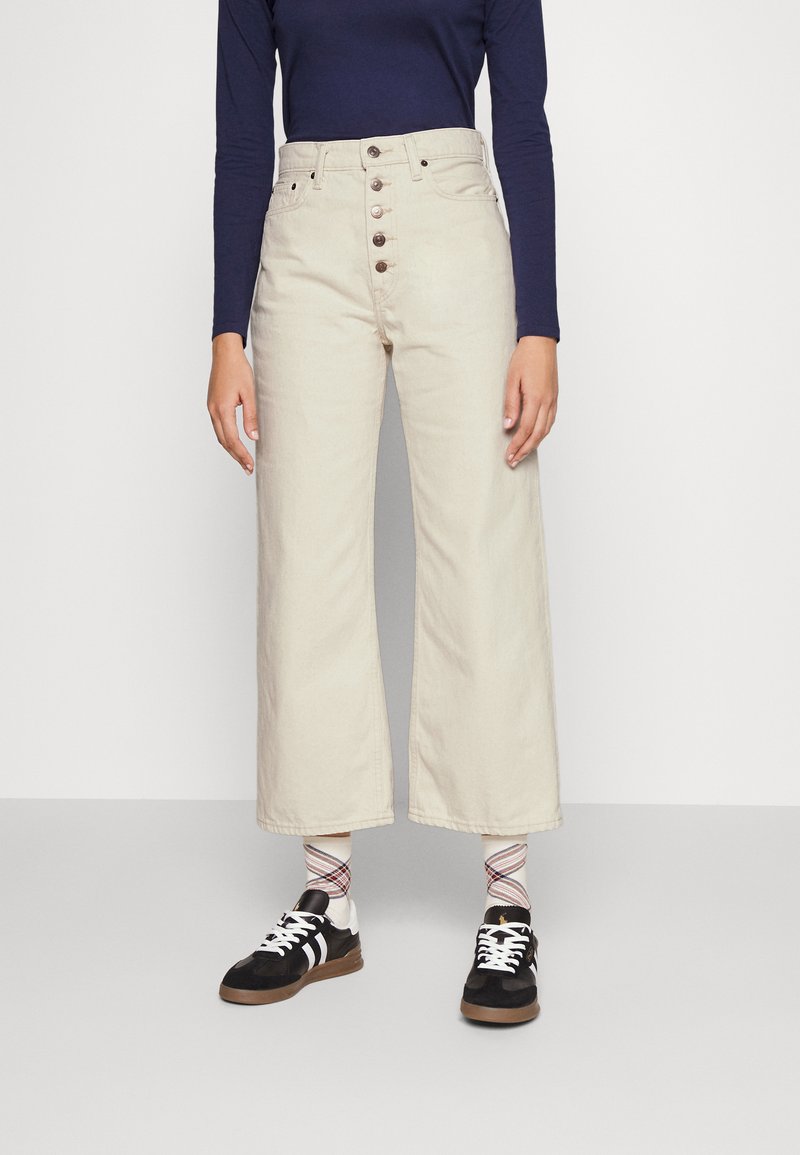 Beżowe dżinsy o szerokich nogawkach z wysokim stanem, zapinane na guziki z przodu, o miękkiej fakturze, połączone z czarnymi trampkami i skarpetkami w wzory.