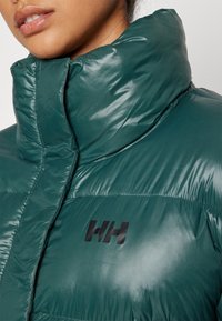 Gros plan d'une personne portant une doudoune vert foncé avec un col montant et un logo noir "HH" sur la poitrine.