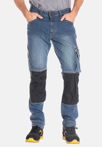 Pantalons cargo en denim avec un lavage bleu clair, des accents en tissu noir aux genoux, plusieurs poches latérales et une poche utilitaire zippée.