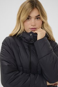 Melnā polsterētā jaka ar augstu apkakli, auklu un sprādzēm. Raksturīga šūta faktūra un mīksts materiāls siltumam.