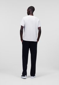 Witte katoenen t-shirt met korte mouwen, gecombineerd met zwart loszittende broek. Model staat met handen in de zakken, met de rug naar voren.