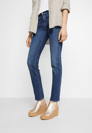 Frau trägt blaue Skinny-Jeans, ein beiges Hemd und goldene Keilsandalen mit geflochtenem Plateauabsatz, stehend vor weißem Hintergrund.