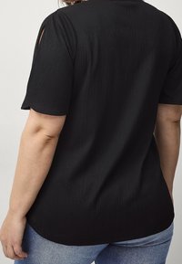 Top corto nero a maniche corte con un tessuto strutturato, caratterizzato da piccoli ritagli sulle spalle e un orlo curvo, indossato con jeans blu.