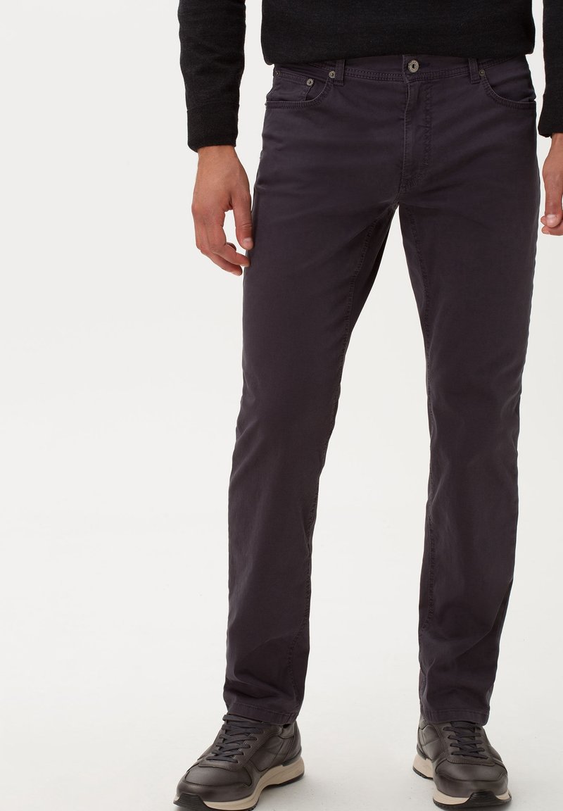 Uomo che indossa pantaloni neri slim-fit, scarpe da ginnastica nere con suole beige e un maglione nero a maniche lunghe, in piedi contro uno sfondo semplice.