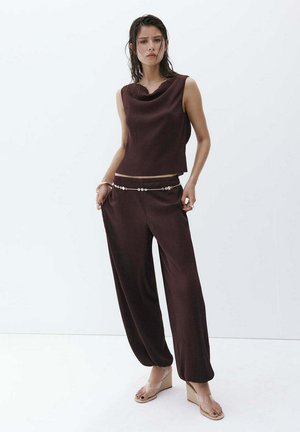 Femme portant un haut sans manches marron foncé et un pantalon large assorti avec une chaîne de taille perlée et des sandales compensées beiges.