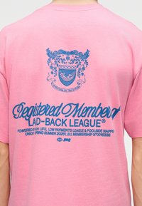 Πίσω μέρος ροζ πουκαμίσου με μπλε έμβλημα που απεικονίζει δύο δράκους και αιώρα, και κείμενο "Εγγεγραμμένο Μέλος της Laid-Back League" μαζί με επιπλέον μικρό κείμενο.