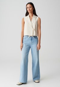 Gilet beige senza maniche, accorciato, con un solo bottone, abbinato a jeans a gamba larga blu chiaro. Tessuto liscio, vestibilità rilassata e vita con cordoncino.