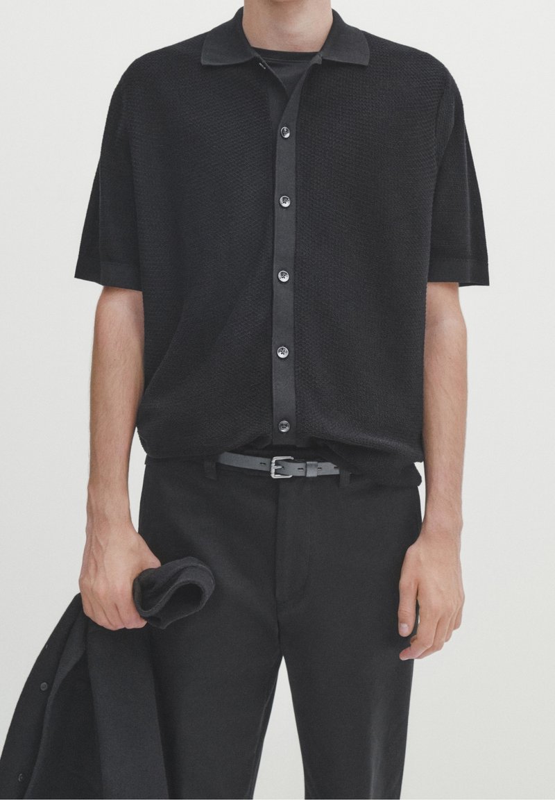 Homme portant une chemise noire à manches courtes boutonnée, rentrée dans un pantalon noir avec une ceinture noire, tenant une veste noire pliée dans sa main gauche.