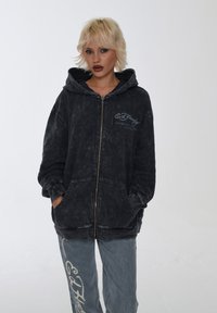 Sort zip-up hoodie med en falmet tekstur, forlommer og broderet logo. Kombineret med lyseblå jeans med iøjnefaldende grafisk tekst.