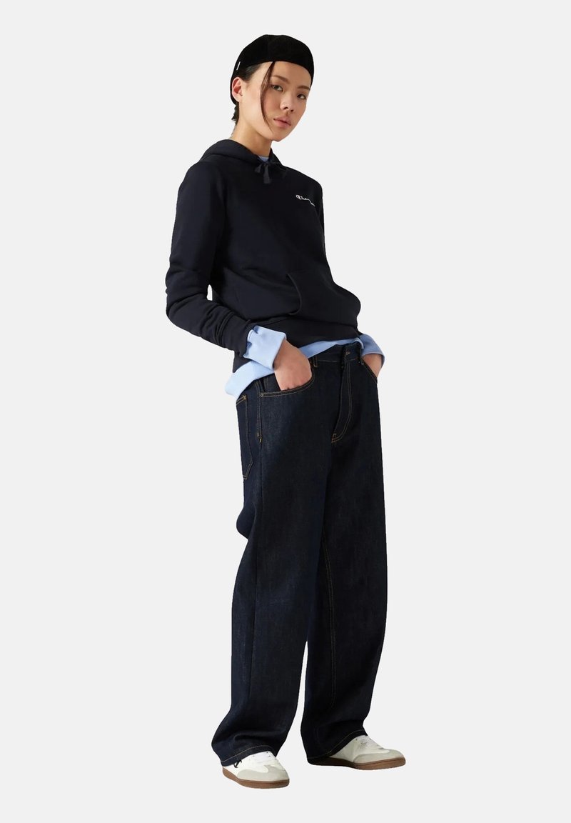 Felpa blu navy con tasca frontale, camicia azzurra chiara sotto e jeans larghi in denim scuro. Sneakers bianche completano l'outfit.