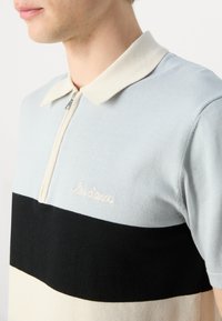 Polo de malha azul claro e preto com mangas curtas, colarinho com fecho éclair, faixa horizontal creme e texto bordado "les deux."