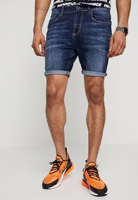 Denim shorts in dark blue with rolled cuffs, featuring a classic five-pocket design. Paired with black and orange athletic shoes.