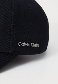 Căciulă de culoare bleumarin, fabricată din material ribbed, cu un logo argintiu "Calvin Klein" în față, având un design structurat și o margine curbată.