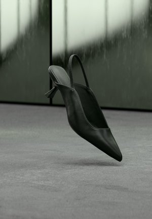 Sort læder slingback højhælet sko med spids tå svævende over gråt tæppe, sløret grønlig væg i baggrunden.