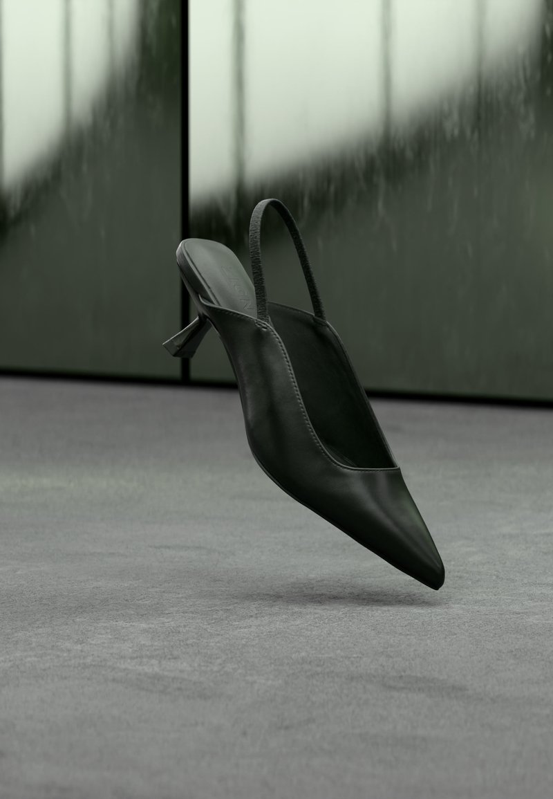 Sort læder slingback højhælet sko med spids tå svævende over gråt tæppe, sløret grønlig væg i baggrunden.