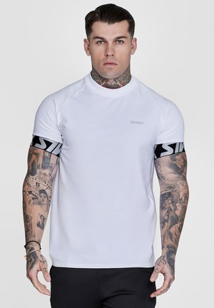 Mann med tatoveringer på nakke og armer iført en hvit kortermet skjorte med svarte logobånd på ermene, stående foran en ensfarget bakgrunn.