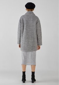 Grå, texturerad oversizejacka med krage, kombinerad med en grå stickad kjol och svarta ankelboots. Bakgrunden är neutral och minimalistisk.