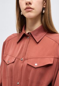 Γυναίκα που φοράει πουκάμισο χρώματος σκουριάς με κουμπιά και τσέπες με καπάκια, καθώς και περλέ κρεμαστά σκουλαρίκια, με ορατά ίσια καστανά μαλλιά.