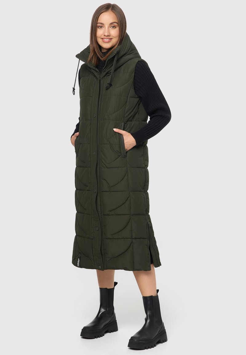 Veste en duvet longue de couleur vert foncé, avec un col haut, une fermeture éclair à l'avant, des poches latérales et une capuche ajustable, associée à des bottes noires imposantes.