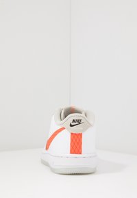 Nike-sneaker med en vit ovandel och orange accenter, texturerad häl och en grå tunga. Gummisula med en strömlinjeformad design.