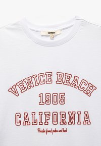 Camiseta de algodón blanca con texto rojo: "VENICE BEACH 1905 CALIFORNIA" con letras curvas, cuello redondo y textura suave.