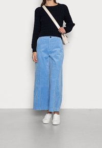 Pantaloni larghi in velluto a coste blu abbinati a un maglione nero a coste e sneaker bianche. L'outfit include una borsa a tracolla di colore chiaro.