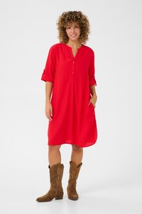 Robe rouge, midi, avec un col en V, des manches trois-quarts, une patte de boutons et des poches latérales, associée à des bottes de cow-boy en suède marron.