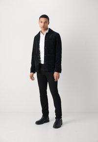 Chaqueta de pana negra con cierre de botones y dos bolsillos frontales, llevada sobre una camisa blanca, combinada con pantalones ajustados negros y botas de tobillo.