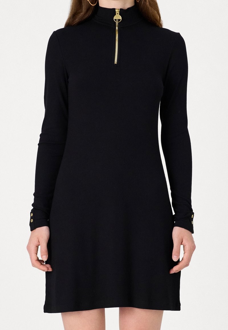 Femme portant une robe noire à manches longues avec une fermeture éclair dorée au cou et des boutons dorés aux poignets, debout devant un fond uni.