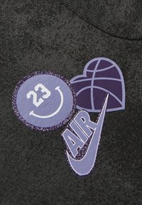 Svart stoff med tre lilla grafiske merker: et sirkulært smil med "23," et hjerteformet basketball, og et swoosh-logo med "AIR."