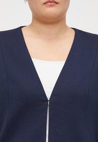Tamno plavi odjevni predmet s V-izrezom, koji ima blago otvorenje sprijeda. Nosite ga preko bijele košulje, s glatkom teksturom tkanine.