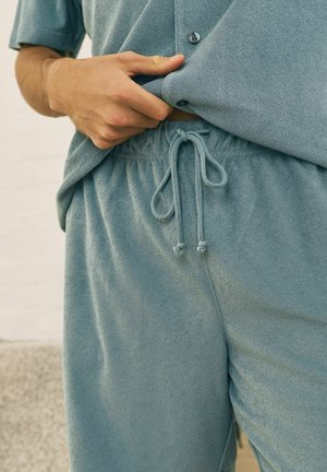 Mann, der blaue Frotteeshorts mit Kordelzug anpasst und ein passendes, geknöpftes Hemd trägt, Nahaufnahme auf Taille und Hand.