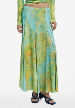 Longue jupe plissée avec motif aquarelle vert et jaune, portée avec des sandales à talons ouvertes bleues et un top court tie-dye vert.