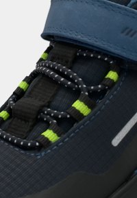 Lähedane kaader tumesinisest kingast, millelt on mustad elastsed paelad, millel on peegeldavad valged täpid ja neoonrohelised lisad, koos sinise Velcro rihmaga.