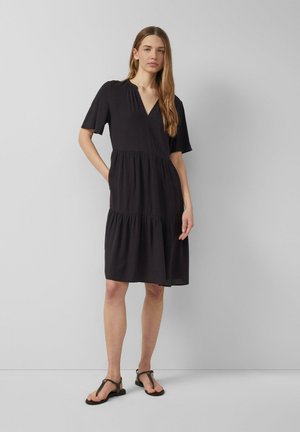 Frau mit langen Haaren, die ein knielanges schwarzes Kleid mit kurzen Ärmeln und schwarzen Sandalen trägt, steht vor einem schlichten, hellgrauen Hintergrund.