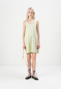 Vestido xadrez verde claro com decote quadrado. Bolsa branca com detalhe trançado. Sapatos planos pretos com atacadores. Estrutura de tecido suave.