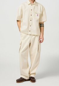 Camisa beige de manga corta con cierre de botones y bolsillo en el pecho; pantalones cargo beige con bolsillos laterales; zapatos marrón oscuro con detalle de costura.