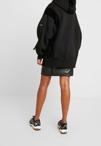 Felpa nera oversize con pannelli laterali texturizzati, abbinata a una minigonna di pelle nera e sneakers sportive nere con dettagli colorati.
