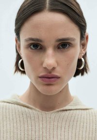 Model z kratkimi, rjavimi lasmi, ki nosi svetlo bež kapuco z rebrasto teksturo. Oznakane lastnosti vključujejo obroče in nevtralen make-up.