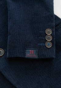 Tumesinine kootud kangas peente soontega, millel on kolm pruuni nööpi ja punane-sinine brändipatch mansetil.