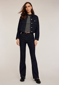 Donker denim cropped jack met knoopsluiting, gecombineerd met hoge, donkere flared jeans. Model draagt een zwart-wit gestreepte top.