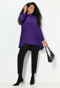 Fialová vrstvená blůza se stojáčkem, s detaily na knoflících, spárovaná s černými slim-fit kalhotami a botami s špičatou špičkou, držící černou kabelku.