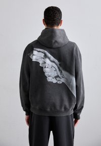 Sudadera gris oscuro con un gran gráfico de una mano en monocromo en la parte de atrás. Tejido suave, diseño con capucha, puños acanalados y dobladillo. Corte casual.