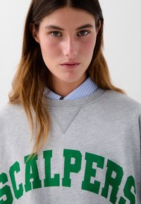 Ung kvinde med langt brunt hår iført en grå sweatshirt med grøn tekst "SCALPERS" og en blåstribet skjorte under.