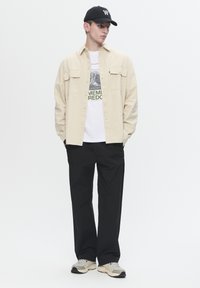 Light beige button-up shirt over a white graphic t-shirt, black trousers, and beige athletic shoes, complemented by a black cap.