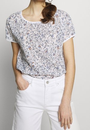 Kobieta w białej bluzce z krótkim rękawem z małym kwiatowym wzorem i białych spodniach z wysokim stanem, z ręką w kieszeni, brązowe włosy związane do tyłu.