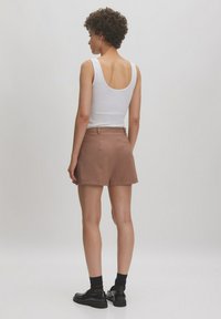 Bruine op maat gemaakte shorts met een gladde textuur en een klassieke snit, gecombineerd met een witte tanktop. Het model staat met de rug naar de camera, waardoor de outfit goed zichtbaar is.