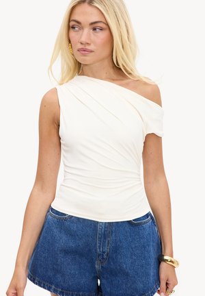 Femme blonde portant un top blanc à une épaule froncé avec un short en jean bleu, boucles d'oreilles dorées, bracelet et bagues à la main gauche.