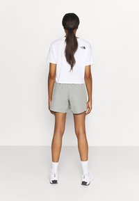 Camiseta blanca recortada con logo negro en la espalda, combinada con pantalones cortos gris claro. El modelo lleva zapatillas blancas y calcetines hasta la pantorrilla.